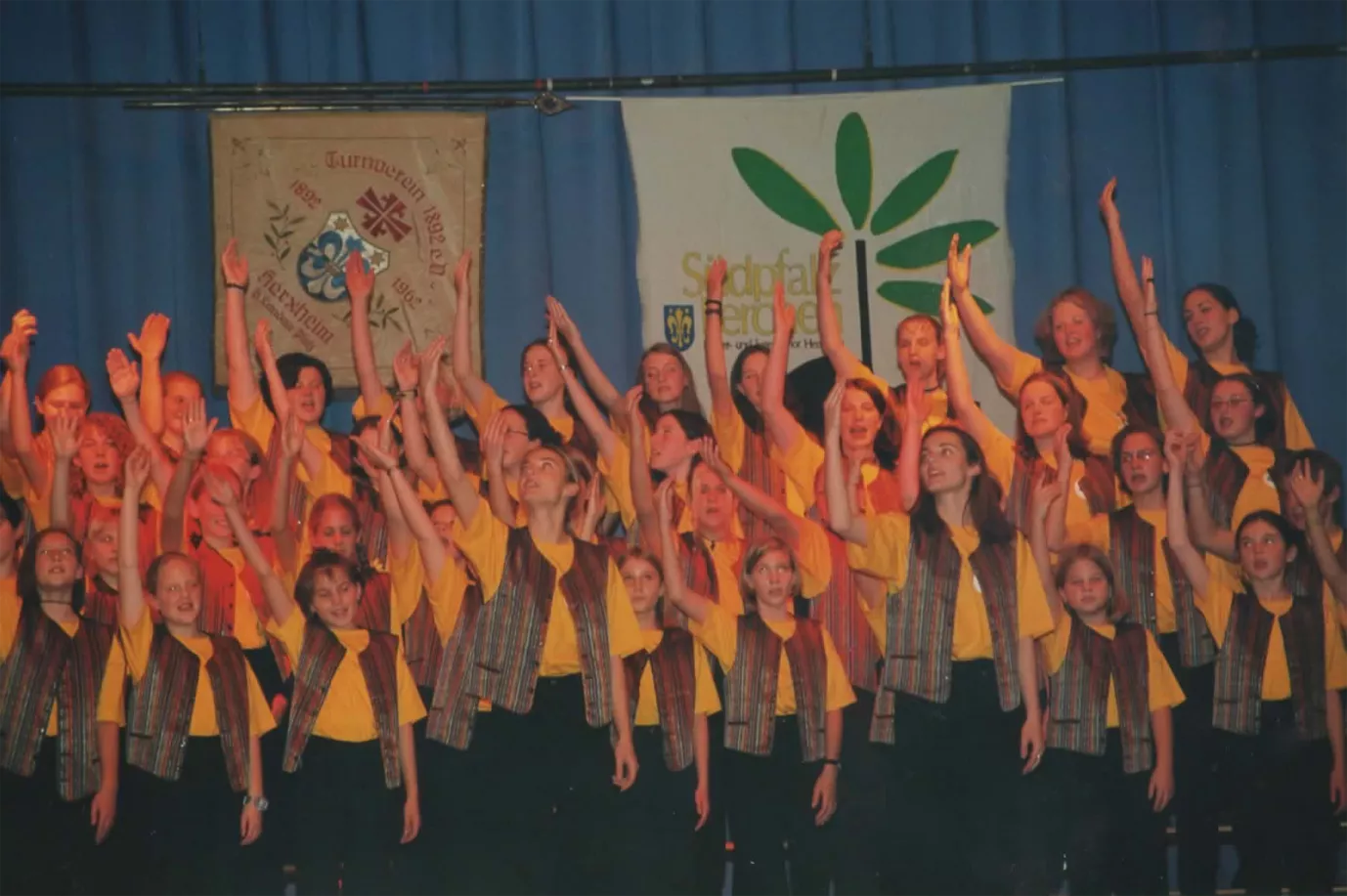 Kinderchor in gelben Hemden mit erhobenen Armen bei einem Konzert vor dem Südpfalz-Lerchen-Banner