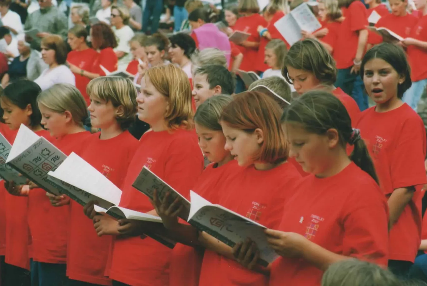Kinderchor in roten Choreley-2000-Shirts singt konzentriert aus Notenheften im Freien