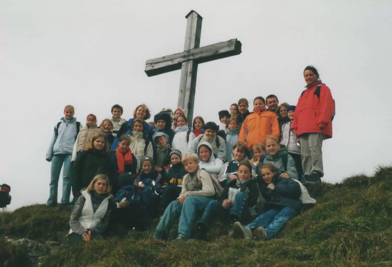 Chorgruppe beim Wandern vor einem großen Gipfelkreuz auf einem nebligen Berg