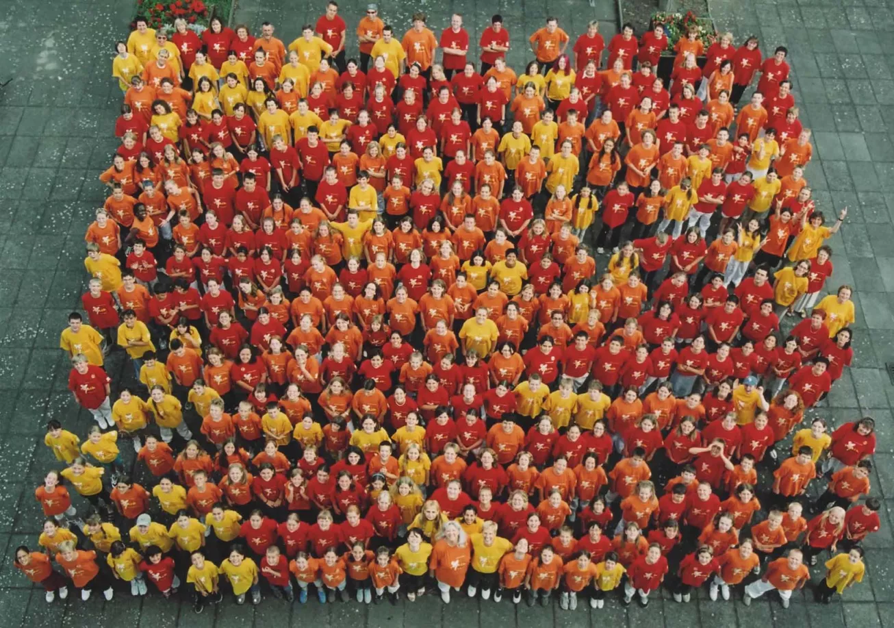 Sehr großes Gruppenfoto aus der Vogelperspektive mit hunderten Sängerinnen und Sängern in roten, orangefarbenen und gelben Shirts