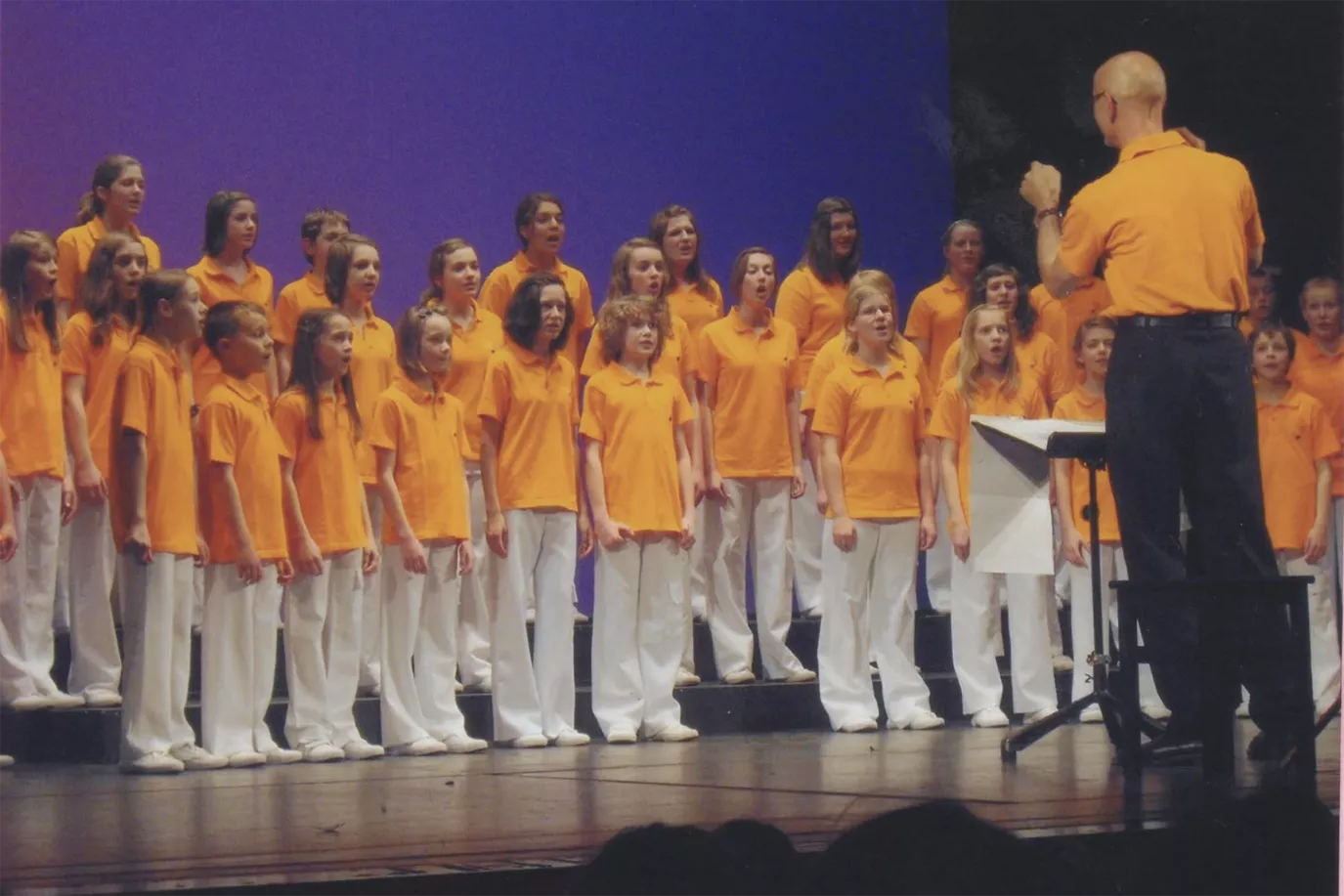 Chor in orangefarbenen Poloshirts singt beim Deutschen Chorwettbewerb mit Dirigent auf der Bühne