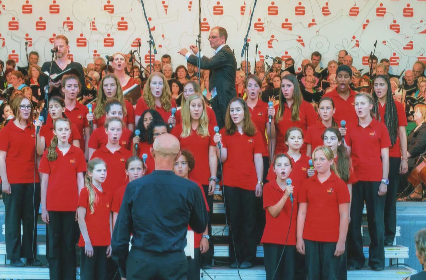 Kinderchor in roten Poloshirts singt mit Mikrofonen bei einem Open-Air-Konzert der Carmina Burana mit Dirigent und Orchester