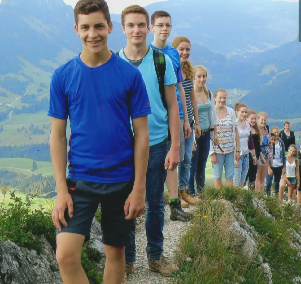 Jugendliche in einer Reihe auf einem sonnigen Bergpfad mit grünem Alpental im Hintergrund