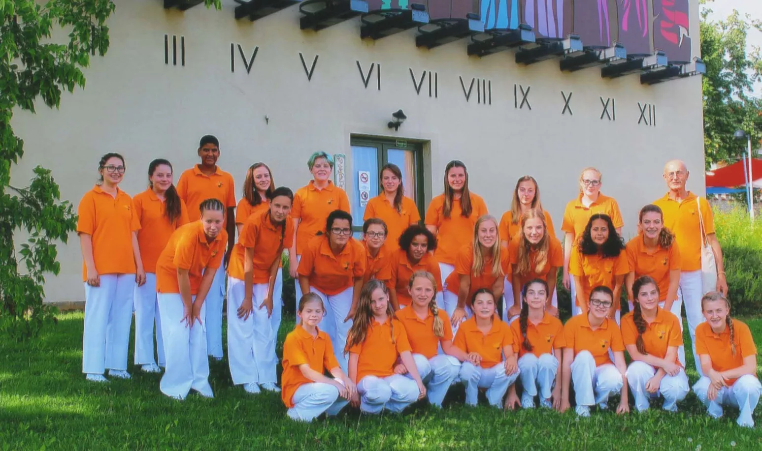 Gruppenfoto des Chors in orangefarbenen Poloshirts vor einer Fassade mit römischen Ziffern