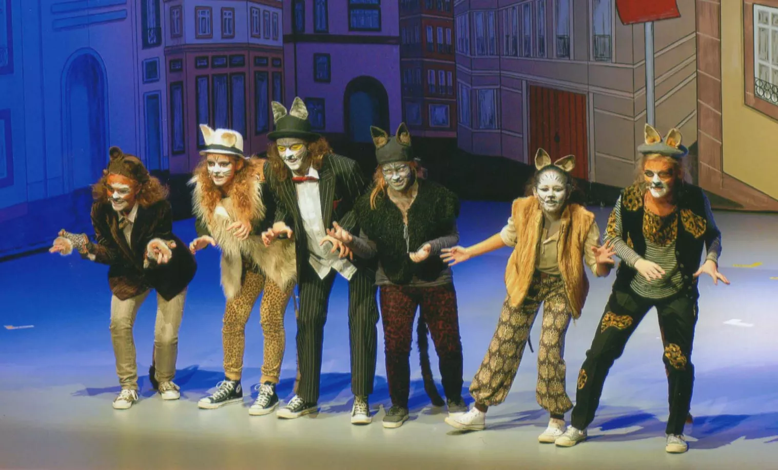 Sechs Kinder in Katzenkostümen und Schminke tanzen vor einer gemalten Stadtkulisse beim Musical Aristocats