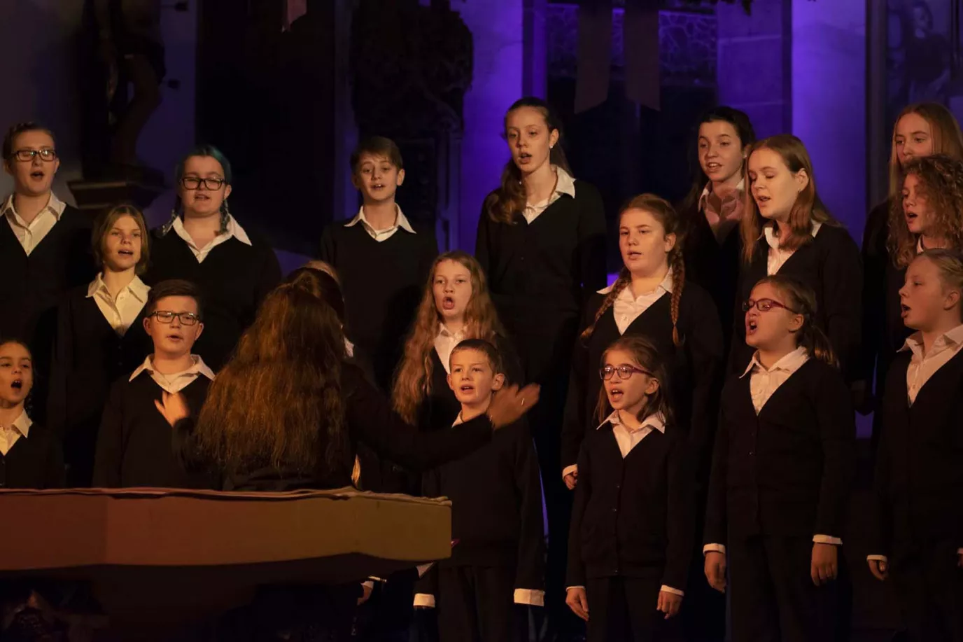 Kinderchor in schwarz-weißer Kleidung singt mit Dirigentin bei einem Weihnachtskonzert im blauen Bühnenlicht