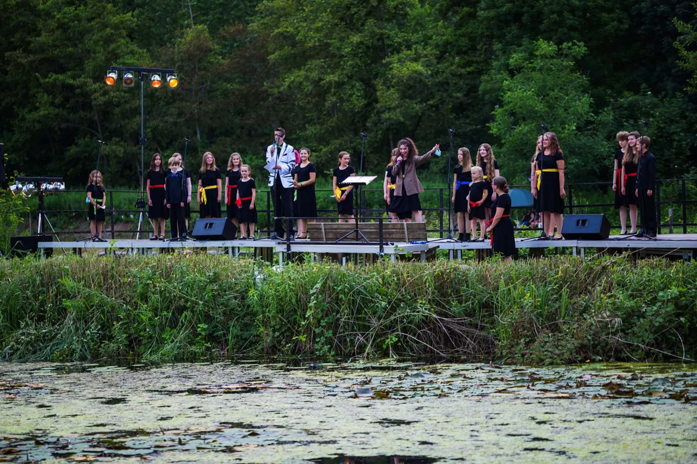 Chor in schwarzer Kleidung singt auf einer Bühne am Seeufer bei einem Open-Air-Konzert in der Dämmerung