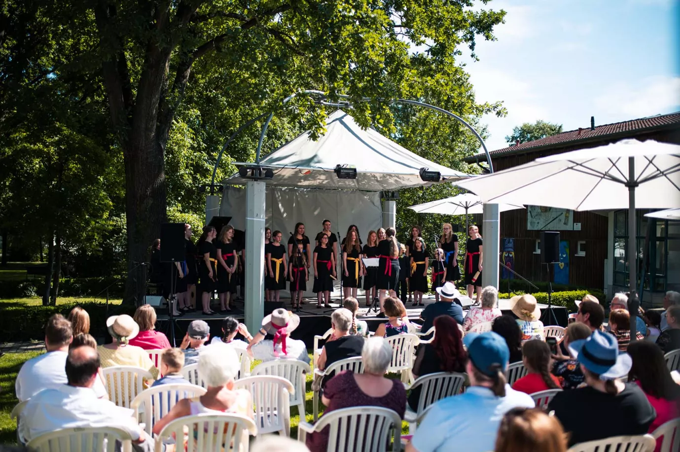 Chor auf einer Pavillonbühne im Park vor sitzendem Publikum bei einem Open-Air-Konzert