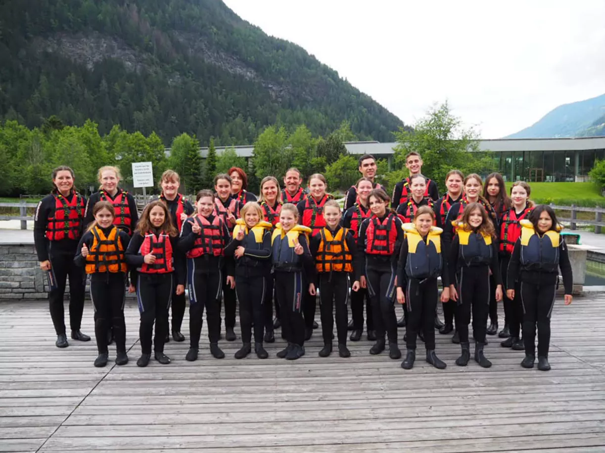 Chorgruppe in Neoprenanzügen und Schwimmwesten posiert auf einem Holzsteg vor Bergpanorama bei den Chorferien in Südtirol