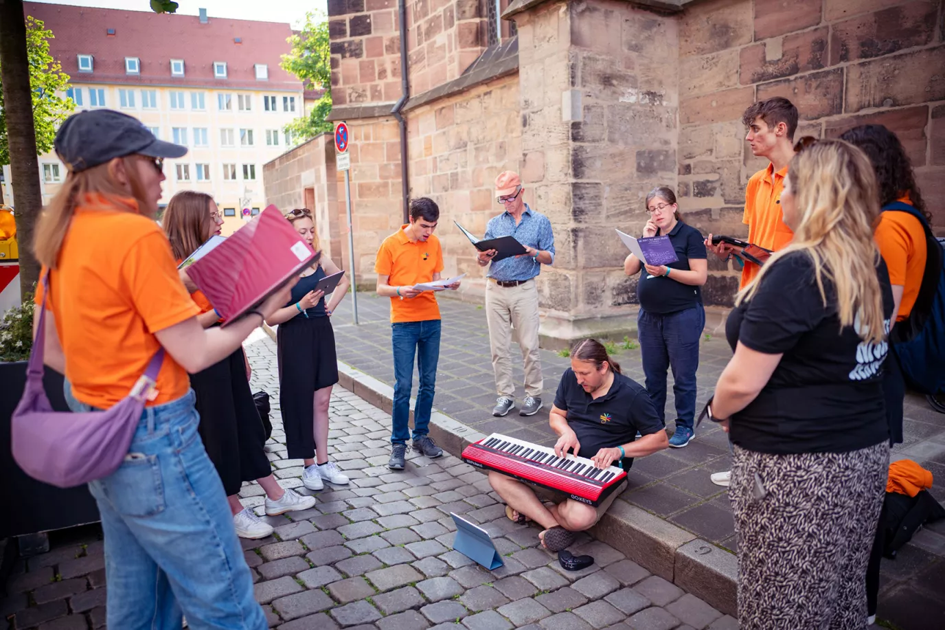 Junge Chorgruppe in orangefarbenen Shirts probt mit Keyboard im Freien an einer Nürnberger Kirche