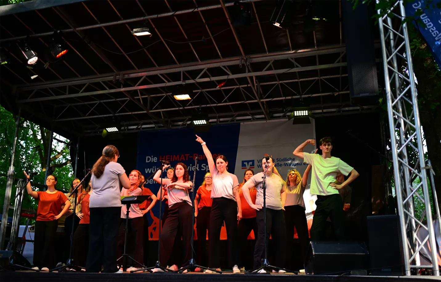 Chor in bunten Sommeroutfits singt und tanzt mit Dirigentin auf einer Freilichtbühne bei Live im Park