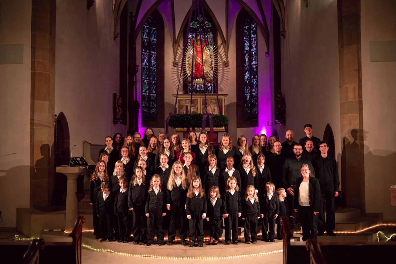 Großer Chor in schwarz-weißer Kleidung mit Dirigentin vor dem beleuchteten Altar beim Adventskonzert in der Kirche